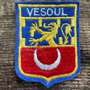 Op de afbeelding: Een rechthoekige geborduurde patch met een blauwe rand en een schildvormig ontwerp. Het bovenste gedeelte is blauw met de tekst VESOUL in het wit en een gele leeuw. Het onderste gedeelte is rood met een witte halve maanvorm in het midden.