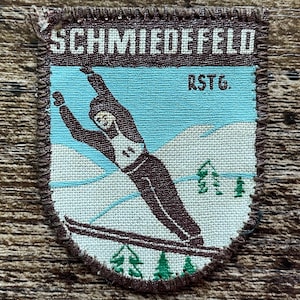 Op de afbeelding: Een schildvormige geborduurde patch met een bruine rand. Het ontwerp toont een skiër in een donkerbruin pak op een lichtblauwe en witte besneeuwde bergachtergrond met groene bomen. De tekst SCHMIEDEFELD staat bovenaan en RSTG. staat rechts.