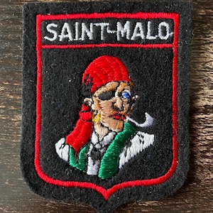 Pode incluir: Um emblema bordado preto em forma de escudo com o texto SAINT-MALO em letras brancas no topo. O centro exibe uma figura bordada colorida usando um lenço vermelho, um tapa-olho e uma peça verde, com um cachimbo branco na boca.
