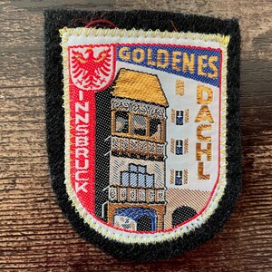 Könnte beinhalten: Ein rechteckiger gestickter Aufnäher mit schwarzem Filzrand. In der Mitte befindet sich eine detaillierte Darstellung des Goldenen Dachls in Innsbruck, Österreich, mit dem Text GOLDENES DACHL und INNSBRUCK sowie einem roten Adleremblem.