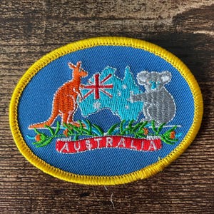 Può includere: Toppa ovale ricamata con bordo giallo, su sfondo blu. Il disegno raffigura un canguro arancione, un koala grigio e una mappa dell'Australia con la bandiera Union Jack. La parola "AUSTRALIA" è in rosso.