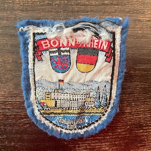 Könnte beinhalten: Ein rechteckiger gestickter Aufnäher mit blauem Rand und der Aufschrift BONN-RHEIN oben. Er zeigt zwei Wappen, eines mit einem roten Löwen und eines mit der deutschen Flagge, über dem Bundeshaus-Gebäude mit der Aufschrift BUNDESHAUS 1981.