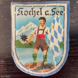 以下が含まれることがあります： Kochel a. Seeという文字が入った水色の縁取りの長方形の布製ワッペン。伝統的な衣装を着た人物が、スパイク付きのメイスと青と白の市松模様の旗を持ち、山を背景に立っている様子が描かれています。