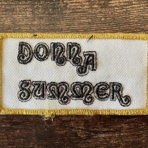 May include: A rectangular patch embroidered with the name "Donna Summer" in black on a white background. The patch has a gold border. The text is in a decorative font.