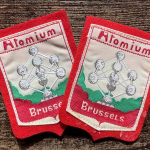 Könnte beinhalten: Zwei rechteckige rote Filzaufnäher mit einer weißen Stickerei des Atomiums. Das Design zeigt den in Rot gestickten Schriftzug Atomium oben und Brussels unten auf einem cremefarbenen Hintergrund mit grünen Akzenten.