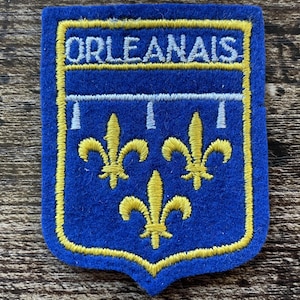Op de afbeelding: Een schildvormige patch van blauw vilt met het woord ORLEANAIS in wit geborduurd aan de bovenzijde. Het ontwerp bevat een witte horizontale balk met drie hangers boven drie in geel geborduurde fleur-de-lis symbolen.