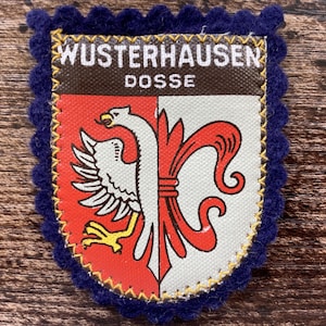Op de afbeelding: Een schildvormige stoffen patch met een donkerblauwe geschulpte vilten rand. Het bovenste gedeelte is bruin met de witte tekst WUSTERHAUSEN DOSSE. Het onderste gedeelte heeft een gespleten ontwerp met een witte adelaar op rood en een rode lelie op wit.