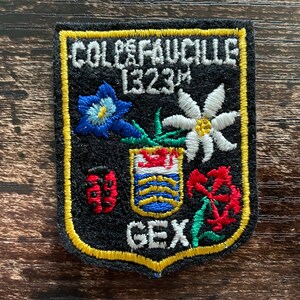 Könnte beinhalten: Ein schwarzer bestickter Aufnäher mit gelbem Rand und dem Text COL DE FAUCILLE 1323M sowie GEX. Das Design zeigt eine blaue Blume, ein weißes Edelweiß, einen roten Marienkäfer, ein zentrales Wappen mit rotem Löwen und rote Blumen auf schwarzem Grund.