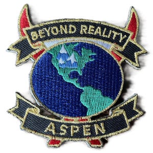 May include: An embroidered patch featuring a blue globe with green continents and white mountain peaks. A black banner above reads BEYOND REALITY in gold, and a black banner below reads ASPEN in gold. The design includes red horn-like accents.