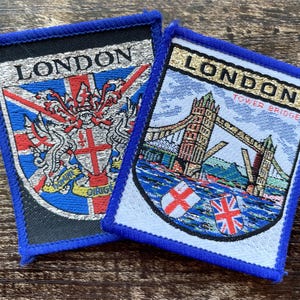 Puede incluir: Dos parches rectangulares bordados con bordes azules. El parche izquierdo presenta la palabra LONDON sobre un escudo y la bandera Union Jack. El parche derecho muestra LONDON y TOWER BRIDGE, representando el puente sobre el agua.