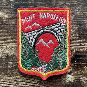 Peut inclure: Un écusson brodé rectangulaire sur fond en feutrine rouge représentant un pont en pierre, des arbres verts et des sommets montagneux blancs. Le texte PONT NAPOLEON est brodé en blanc en haut. L'écusson est bordé d'un fin liseré jaune.