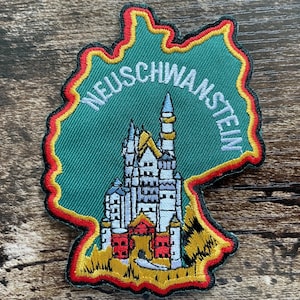 May include: An embroidered patch shaped like the map of Germany with a green background and a red, yellow, and black border. It features a detailed white and grey castle with red accents and the word NEUSCHWANSTEIN in white capital letters.