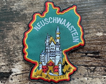 May include: An embroidered patch shaped like the map of Germany with a green background, a red and yellow border, and the word NEUSCHWANSTEIN in white text. The center features a detailed depiction of Neuschwanstein Castle.