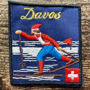 Può includere: Una toppa ricamata rettangolare con sfondo blu che presenta la parola Davos in caratteri gialli. Il design raffigura uno sciatore con un top rosso, un gilet bianco e pantaloni blu, con una bandiera svizzera rossa nell'angolo e neve bianca.