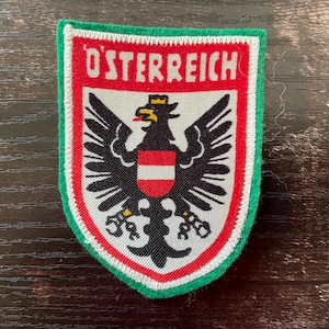 Könnte beinhalten: Ein schildförmiger gestickter Aufnäher mit dem Schriftzug O'STERREICH in Weiß auf rotem Grund. In der Mitte befindet sich ein schwarzer Adler mit goldener Krone, einem rot-weißen Schild auf der Brust und gesprengten Ketten an den Füßen, grün umrandet.