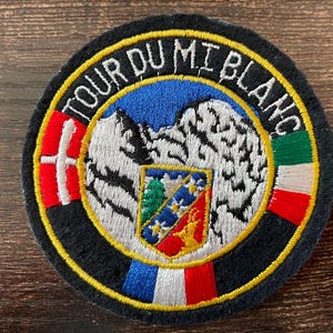 Könnte beinhalten: Runder, gestickter Aufnäher mit dem Text "TOUR DU MI BLANC". Das Design umfasst eine Berglandschaft, Flaggen und ein Wappen. Der Rand ist schwarz mit gelber Kontur.