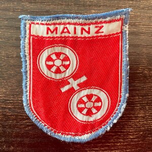 May include: A shield-shaped embroidered patch featuring a red background with two white wheels and a white cross symbol. The word MAINZ is stitched in red letters on a white horizontal band at the top. The patch has a light blue stitched border.