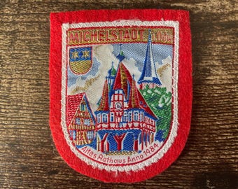 May include: A red felt patch featuring an embroidered scene of the Michelstadt town hall. The design includes the text MICHELSTADT and Altes Rathaus Anno 1484, a small shield with two stars, and a detailed depiction of the historic building.