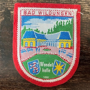Peut inclure: Un écusson rectangulaire en feutrine rouge représentant une scène brodée d'un bâtiment jaune au toit bleu à Bad Wildungen. Sous le bâtiment, une zone verte affiche deux petits écussons, l'un avec un lion et l'autre avec une étoile, et le texte Wandelhalle.