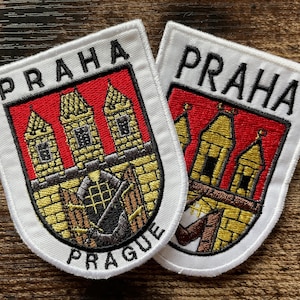 Op de afbeelding: Twee geborduurde patches met het wapen van Praag. Elke schildvormige patch toont een rode achtergrond met drie gouden torens, een stenen muur en een open poort. De tekst PRAHA is in het zwart bovenaan geborduurd en PRAGUE onderaan.