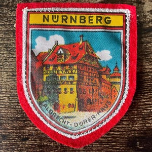 Peut inclure: Un écusson en tissu en forme de bouclier avec une bordure rouge présentant une illustration imprimée de la maison d'Albrecht Dürer à Nuremberg. La bannière jaune supérieure affiche le texte NURNBERG et le texte incurvé en bas indique ALBRECHT-DURER-HAUS.