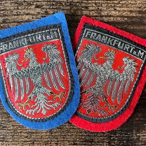 Puede incluir: Dos parches bordados en forma de escudo con un águila plateada sobre fondo rojo. Un parche tiene un borde de fieltro azul y el otro un borde de fieltro rojo. Ambos parches muestran el texto FRANKFURT a.M en letras plateadas arriba.