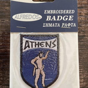 Op de afbeelding: Een geborduurde schildvormige patch met een blauwe achtergrond, een lichtgekleurde figuur en het woord ATHENS in witte letters. De patch zit in een doorzichtige plastic verpakking met een kopkaart met het opschrift ALFREDOS EMBROIDERED BADGE.