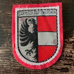 Könnte beinhalten: Rechteckiger Aufnäher mit rotem Rand und dem Text "GARMISCH-PARTENKIRCHEN" oben. Der Aufnäher zeigt ein Schilddesign mit einem schwarzen Adler auf silbernem Hintergrund auf der linken Seite und einem rot-weiß karierten Muster auf der rechten Seite.