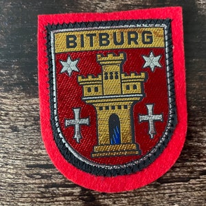 Könnte beinhalten: Ein rot-goldener, gestickter Aufnäher mit dem Wort "BITBURG". Das Design zeigt eine Burg, Sterne und Kreuze. Er hat einen roten Rand mit schwarzem und silbernem Besatz. Wahrscheinlich ein Abzeichen oder Emblem.