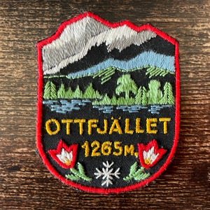 Op de afbeelding: Een zwarte geborduurde patch met een berglandschap met een meer en bos. De tekst luidt OTTFJÄLLET 1265M. Het ontwerp bevat twee rood-witte bloemen aan de onderkant, een wit sneeuwvloksymbool en een felrode randafwerking.