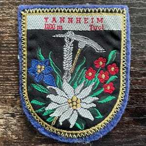 Könnte beinhalten: Ein schildförmiger, bestickter Aufnäher mit blauem Rand. Der obere Bereich zeigt den Text TANNHEIM 1100 m Tirol. In der schwarzen Mitte befinden sich eine weiße Edelweißblüte, ein blauer Enzian, rote Blüten, grüne Blätter und ein silberner Eispickel.
