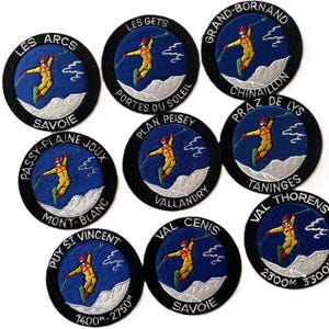May include: A collection of nine circular embroidered patches featuring a skier in a yellow suit against a blue sky and white mountains. Each patch displays a unique French ski resort name in white text on a black border, including Les Arcs, Les Gets, Grand-Bornand, Passy-Plaine Joux, Plan Peisey, Praz de Lys, Puy St Vincent, Val Cenis, and Val Thorens.