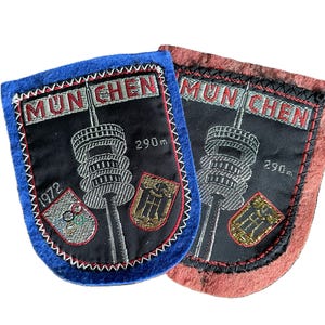 Könnte beinhalten: Zwei gestickte Aufnäher mit der Aufschrift MUN CHEN und 290m. Das Design zeigt einen hohen Turm, die Jahreszahl 1972 und zwei Wappen. Ein Aufnäher hat einen blauen Rand, der andere einen rotbraunen Rand. Beide haben schwarze Mitten mit weißer Stickerei.