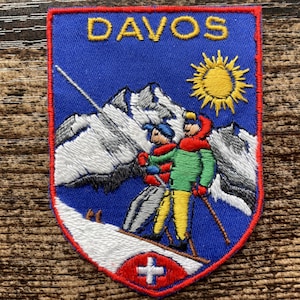 Op de afbeelding: Een rechthoekige geborduurde patch met een blauwe achtergrond en de gele tekst DAVOS bovenaan. Het ontwerp toont twee skiërs in groene en gele kleding tegen besneeuwde bergen, een gele zon en een rood embleem met een Zwitsers kruis.