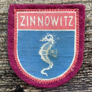 Puede incluir: Un parche de tela rectangular con un borde de fieltro magenta oscuro. La sección superior presenta una banda horizontal rosa con el texto blanco ZINNOWITZ. La sección inferior es un campo azul que muestra un gráfico de caballito de mar.