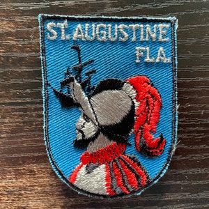 May include: Embroidered patch with the text "ST. AUGUSTINE FLA." above a depiction of a conquistador in profile, wearing a red and black plumed hat. The patch has a blue background and a black border.
