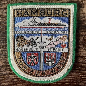 Op de afbeelding: Een schildvormige geborduurde patch met de tekst HAMBURG en TOR ZUR WELT. Het toont het schip TS HAMBURG, een leeuw met het label HAGENBECK, een figuur met het label ST. PAULI en twee wapenschilden, alles omrand door een groene vilten rand.