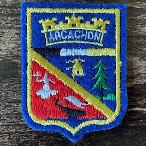 Peut inclure: Un écusson brodé en forme de blason avec un fond bleu et une bordure jaune. En haut, une couronne murale jaune surmonte le texte ARCACHON. La partie inférieure est divisée en diagonale, montrant une silhouette blanche, une cloche jaune, un pin vert et un bateau noir sur un champ rouge et bleu.