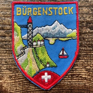 Può includere: Una toppa ricamata a forma di scudo con sfondo blu e bordo rosso. Presenta il testo giallo BÜRGENSTOCK sopra un paesaggio di scogliere verdi, una torre alta, montagne bianche, un lago blu, una barca a vela e una croce svizzera bianca.