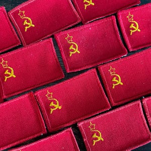 May include: A collection of red rectangular patches, each embroidered with a gold hammer and sickle symbol topped with a star. The patches are arranged closely together, showcasing the repeating design.