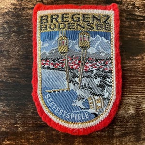 Könnte beinhalten: Gestickter Aufnäher mit rotem Rand. Der Aufnäher zeigt den Text "BREGENZ BODENSEE" und "SEEFEESTSPIELE". Er zeigt eine Berglandschaft mit Seilbahnen und einem See in Blau, Weiß, Gold und Schwarz.