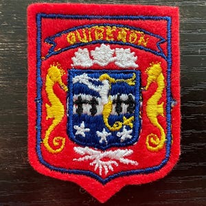 May include: A rectangular red felt patch featuring a blue shield with a white crest, two yellow seahorses, and white stars. The word QUIBERON is embroidered in yellow on a red banner at the top. The design includes white and black details.