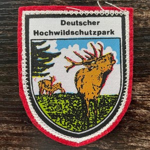 Puede incluir: Un parche bordado en forma de escudo con un borde de fieltro rojo y un borde cosido en blanco. El centro presenta un gráfico de un ciervo y dos ciervos más pequeños en un campo verde bajo un cielo azul, con el texto Deutscher Hochwildschutzpark.