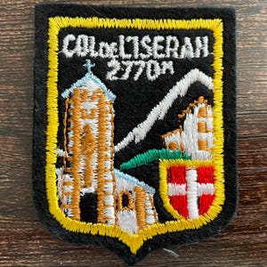 Puede incluir: Un parche bordado rectangular con fondo negro y borde amarillo. Presenta una montaña blanca, una iglesia marrón con techo azul y un escudo rojo con una cruz blanca. El texto dice COL DE L'ISERAN 2770M en hilo de color blanco.