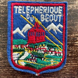 May include: A rectangular embroidered patch with a blue background and a red border. It features white text reading TELEPHERIQUE DU BEOUT above a red cable car, white mountain peaks, yellow cables, and a green landscape with small buildings.