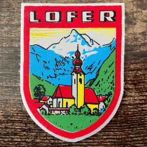 Op de afbeelding: Een schildvormige geborduurde patch met de tekst LOFER in zwarte letters op een rode achtergrond. Het ontwerp toont een gele kerk met een rood dak, groene heuvels en blauwe bergen op de achtergrond, omringd door een dikke rode rand.