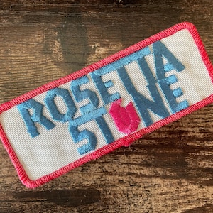 Op de afbeelding: Rechthoekige geborduurde patch met de tekst "ROSETTA STONE" in blauw, met een roze tulpafbeelding. De patch heeft een witte achtergrond en een rode rand.