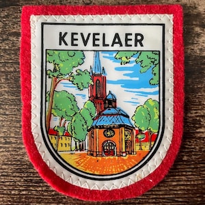May include: A shield-shaped patch with a red felt border and a white stitched edge. The center features a colorful illustration of a church building with a tall steeple, green trees, and a blue sky. The word KEVELAER is printed in black at the top.
