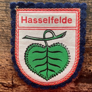 Op de afbeelding: Een stoffen patch met het woord "Hasselfelde" in rode letters. De patch heeft een groen bladerontwerp en is omzoomd met een marineblauwe geschulpte rand. De patch is gestikt met rood en geel garen.