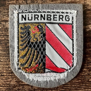 May include: A patch featuring the word "NÜRNBERG" at the top. The patch has a shield design with a black and gold eagle on the left and red and white stripes on the right. The patch is bordered by gray fabric and white stitching.